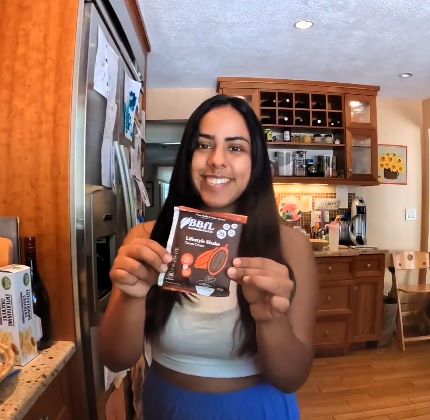 A smiling woman holding up a BBFL Lifestyle Shake packet in a kitchen