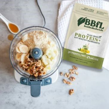 A food processor filled with bananas, walnuts, and powder sits next to a bag of BBFL Plant-Based Protein