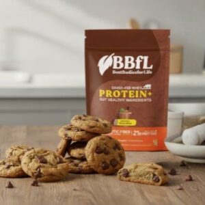 A brown bag of BBFL Grass-Fed Whey Protein+ stands behind a pile of chocolate chip cookies on a wooden table.