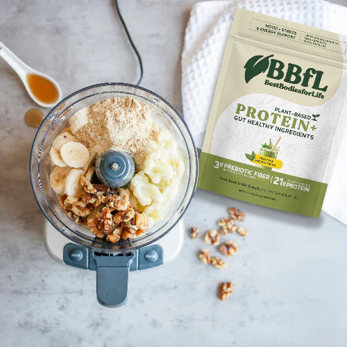 A food processor filled with bananas, walnuts, and powder sits next to a bag of BBFL Plant-Based Protein