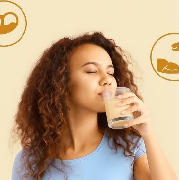 A young woman, wearing a light blue top, sips from a glass of a light-colored beverage with her eyes closed.