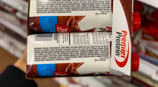 A person's hand holds a multi-pack of Premier Protein chocolate shakes, showing the nutritional information and branding.