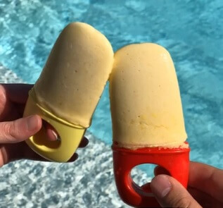 Two hands hold mango popsicles, clinking them together over a bright blue swimming pool.