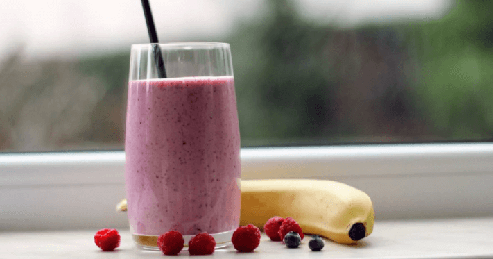 A glass of pink berry smoothie with a straw sits on a windowsill next to raspberries, blueberries, and a banana.
