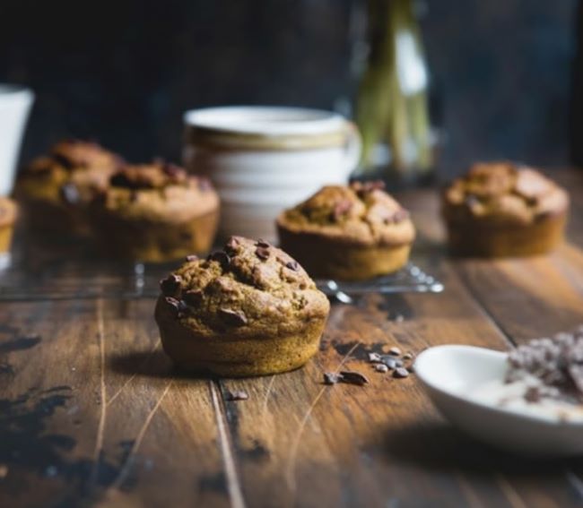 Chocolate chip muffins are arranged on a wooden table, with one in the foreground, and other blurred muffins and kitchen items in the background.