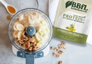 A food processor filled with bananas, walnuts, and powder sits next to a bag of BBFL Plant-Based Protein