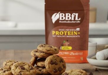 A brown bag of BBFL Grass-Fed Whey Protein+ stands behind a pile of chocolate chip cookies on a wooden table.