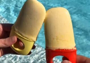Two hands hold mango popsicles, clinking them together over a bright blue swimming pool.