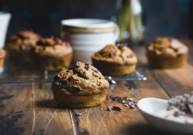 Chocolate chip muffins are arranged on a wooden table, with one in the foreground, and other blurred muffins and kitchen items in the background.