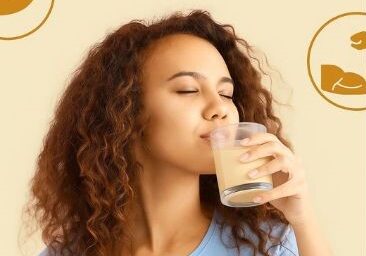 A young woman, wearing a light blue top, sips from a glass of a light-colored beverage with her eyes closed.